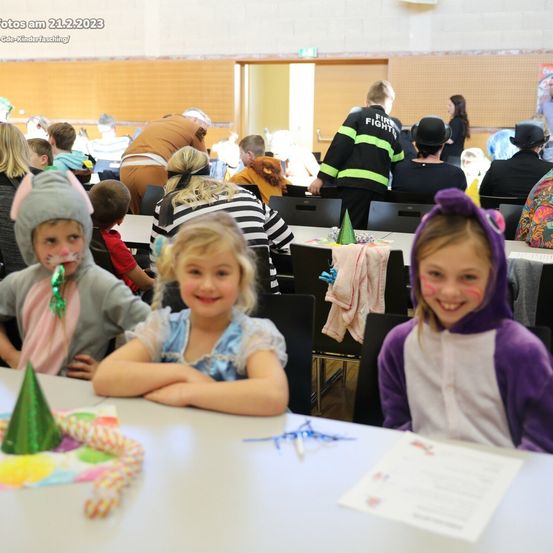 Eine Gruppe von Kindern in Kostümen sitzt lächelnd am Tisch. Ein Mädchen hat Gesichtsbemalung. Hinter ihnen sitzen andere Kinder und Erwachsene, einige in Kostümen, in einem Raum. Ein Feuerwehrmann und eine Person mit Hut sind sichtbar.