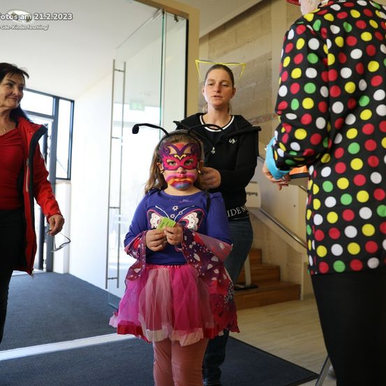 Ein kleines Mädchen in einem Schmetterlingskostüm und einer Maske steht in einem Flur. Eine erwachsene Frau hilft ihr. Zwei andere Frauen sind im Hintergrund.