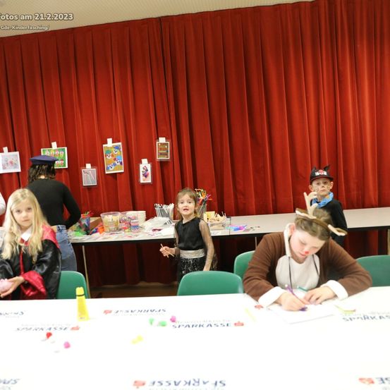 Kinder in Kostümen nehmen an einer Bastelaktivität teil, sitzen um einen Tisch mit verschiedenen Utensilien, vor einem roten Vorhang als Hintergrund.