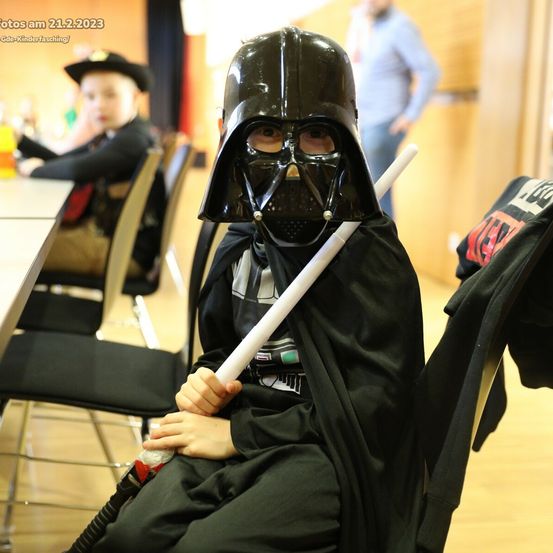 Ein Kind in Darth-Vader-Kostüm sitzt auf einem Stuhl und hält ein Lichtschwert. Ein weiteres Kind und ein Mann sind im Hintergrund. Datumsstempel: 21.2.2023.