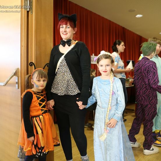 Eine Frau mit Katzenohren und einem Leopardenmuster-Top hält Händchen mit zwei Mädchen, eines in einem Schmetterlingskostüm und das andere in einem blauen Kleid. Sie stehen vor einer Holztür. Dahinter sind weitere Menschen in Kostümen zu sehen.