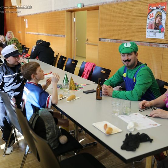 Eine Gruppe von Kindern und Erwachsenen sitzt an einem Tisch mit Essen und Getränken. Ein Kind bläst in ein Pfeifen. Ein Mann in einem grünen Mario-Kostüm sitzt neben einer Frau. Das Foto ist vom 21.2.2023.