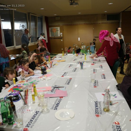 Ein Raum mit Kindern, die Kunsthandwerk machen. Eine Frau hilft ihnen. Es gibt mehrere Flaschen und Papiere auf dem Tisch. Eine Frau mit einer rosa Perücke schaut die Kinder an. Ein Schild steht für Sparkasse.