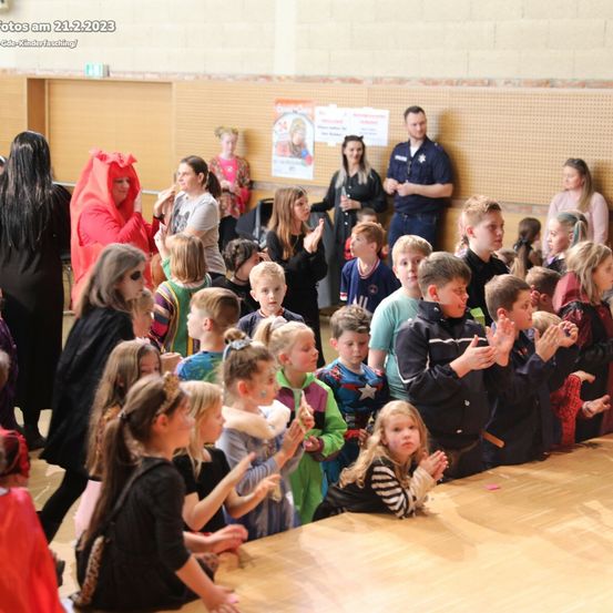 Eine Gruppe von Kindern in verschiedenen Kostümen klatscht und schaut sich eine Aufführung in einer Halle an. Das Datum des Fotos ist 21.2.2023.