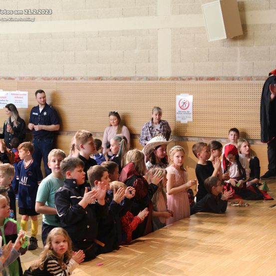 Eine Gruppe von Kindern klatscht mit den Händen, während sie auf einem Holzboden sitzen, wahrscheinlich in einem Saal. Hinter ihnen stehen mehrere Erwachsene, darunter ein Mann in Polizeiuniform. An der Wand sind auch Plakate angebracht. Das Bild hat einen Wasserstempel mit Datum und dem Text 'Kinderfasching'.