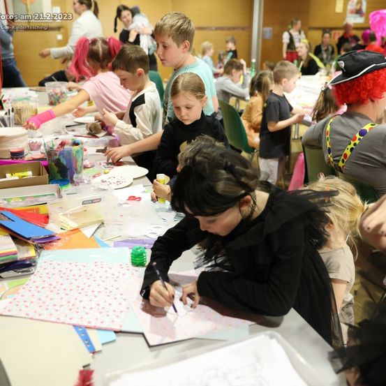 Eine Gruppe von Kindern und Erwachsenen nimmt an einer Bastelaktivität teil. Verschiedene Bastelmaterialien liegen auf dem Tisch. Eine Frau in Kostüm schreibt auf ein Papier. Das Bild hat einen Wasserzeichen mit dem Datum 21.2.2023.