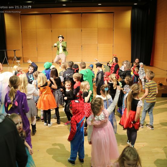 Eine Gruppe von Kindern und Erwachsenen in Kostümen steht auf der Bühne, ein Mann in Clownskostüm führt vor. Die Bühne ist aus Holz mit schwarzem Hintergrund. Datumsstempel lautet 21.2.2023.