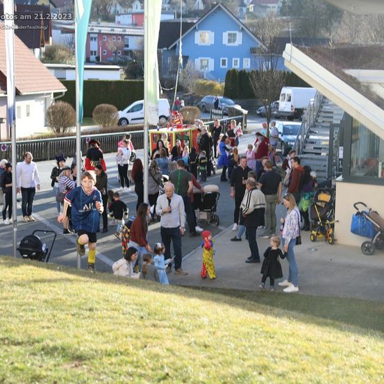 Eine lebhafte Straßenszene am 21. Februar 2023 mit Familien, Kindern in Kostümen und einem Karnevalswagen. Die Straße ist voller Aktivität, mit Menschen, die gehen, stehen und miteinander interagieren.