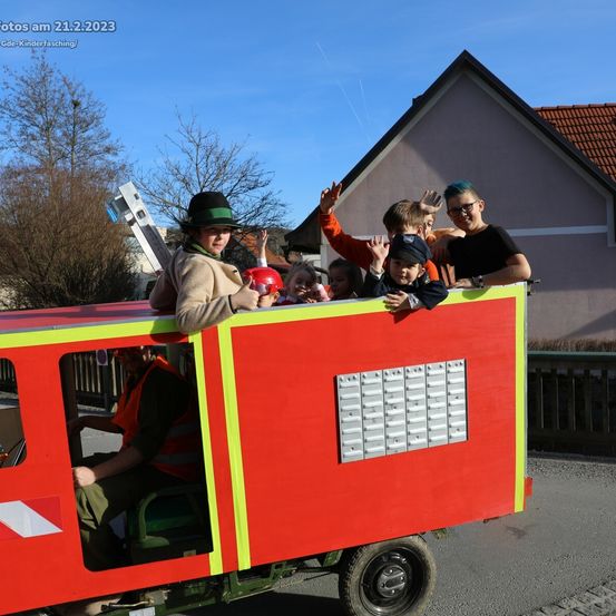 Eine Gruppe von Kindern und Erwachsenen fährt auf einem roten Feuerwehrwagen bei einer Parade, mit einem Mann am Steuer und winkt im Hintergrund.