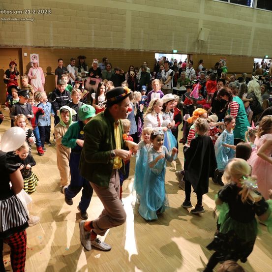 Eine Menschenmenge, darunter Kinder in Kostümen, tanzt am 21. Februar 2023 in einer großen Halle. Ein Mann in einem Hut führt die Kinder an, während andere zuschauen und die Festlichkeiten genießen.
