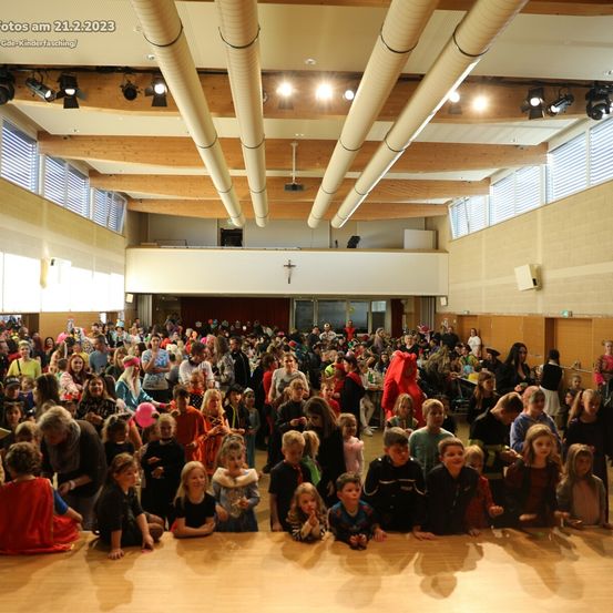 Eine große Menschenmenge in Kostümen versammelt sich in einem Saal, einige sitzend, andere stehend, mit einer Holzdecke und weißen Wänden, aufgenommen am 21.2.2023.