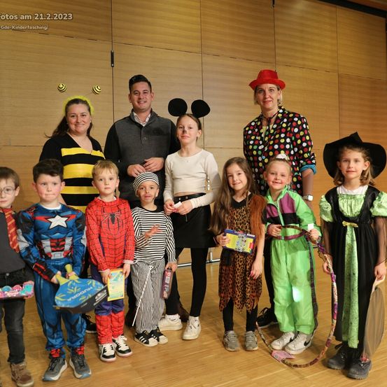 Gruppenfoto von Menschen in Kostümen in einer Halle, datiert auf den 21.2.2023. Die Kostüme umfassen Spiderman, Captain America und verschiedene Tierthemen. Eine Frau in einem gepunkteten Hemd und Hut steht hinter der Gruppe.