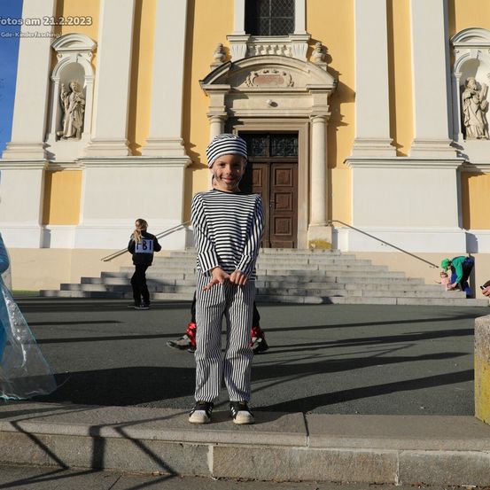 Ein Kind in einem gestreiften Outfit steht am 21. Februar 2023 vor einer Kirche. Hinter dem Kind stehen zwei Personen auf den Stufen, eine in einem Kostüm.