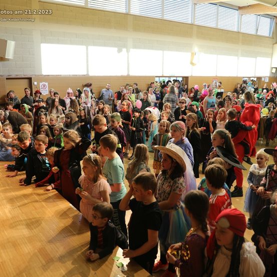 Eine große Menschenmenge von Kindern in verschiedenen Kostümen versammelt sich in einer Halle, einige sitzen auf dem Boden und andere stehen, alle schauen nach vorne, möglicherweise bei einer Halloween-Veranstaltung.