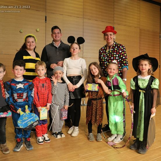 Eine Gruppe von Kindern und Erwachsenen posiert für ein Foto in einem Raum. Sie sind alle in Kostümen gekleidet, einige mit Hüten. Eine Frau trägt ein Punkthemd.