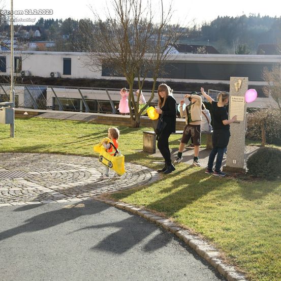 Eine Gruppe von Menschen draußen mit einem Kind in einem gelben Kostüm. Eine Frau hält einen gelben Luftballon. Ein Schild ist in der Nähe und ein Gebäude im Hintergrund.