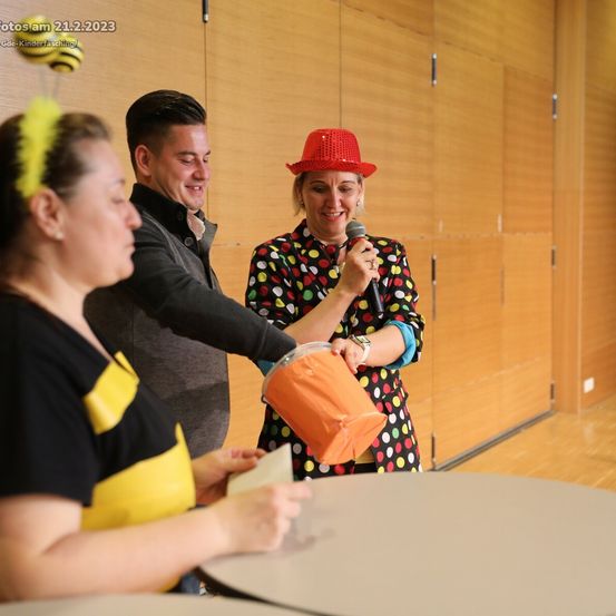 Drei Personen versammeln sich in einem Raum, zwei Frauen und ein Mann. Eine Frau hält ein Mikrofon und einen Eimer und lächelt den Mann an, der ein Stück Papier in der Hand hat. Hinter ihnen steht eine Holzwand.