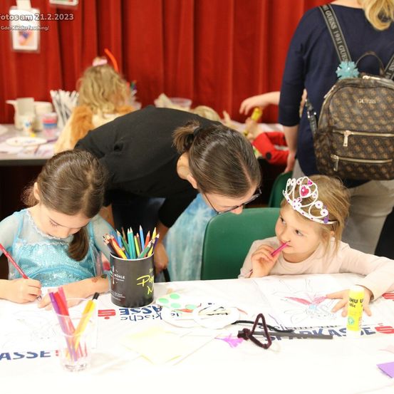 Eine Gruppe von Kindern und einer erwachsenen Frau sind an einem Tisch. Sie beschäftigen sich mit Malaktivitäten. Auf dem Tisch liegen verschiedene Malwerkzeuge und Bastelutensilien. Die Kinder tragen Kostüme, und eines von ihnen trägt eine Krone.