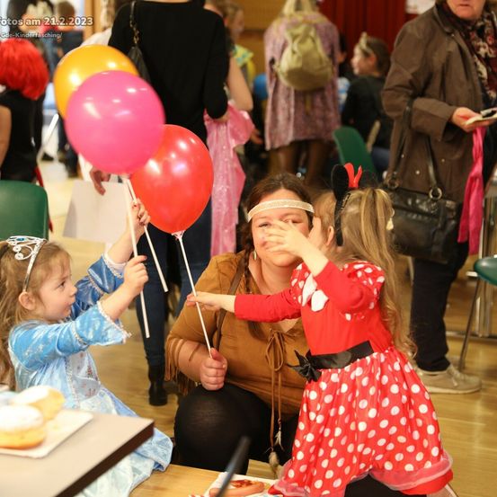 Zwei junge Mädchen in Kostümen halten Luftballons. Eine Frau beobachtet sie lächelnd. Hinter ihnen sind Menschen in verschiedenen Outfits anwesend. Eine Person steht und schaut ein Buch an.