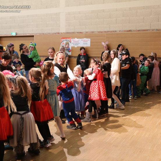 Eine Gruppe von Kindern in Kostümen ist in einer Halle versammelt. Einige tanzen, während andere zuschauen. Eine Frau in einem Kostüm trägt ein Baby.