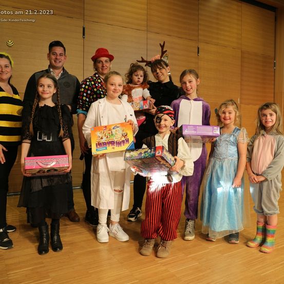 Eine Gruppe von Menschen, darunter Kinder in Kostümen, posiert für ein Foto mit Spielzeug. Ein Mann in Anzug und eine Frau in einer Bienenkostüm stehen an den Seiten. Die Kinder halten Boxen und lächeln.