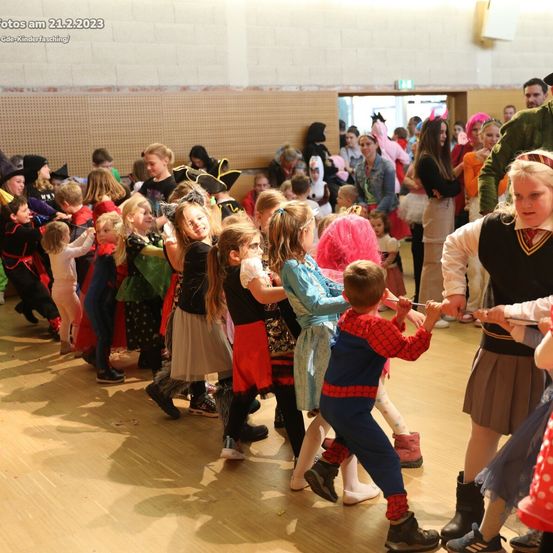 Eine Gruppe von Kindern in verschiedenen Kostümen, darunter Superhelden und Piraten, nehmen an einer Kostümparty teil. Sie tanzen in einer Reihe in einem Raum mit Holzböden.