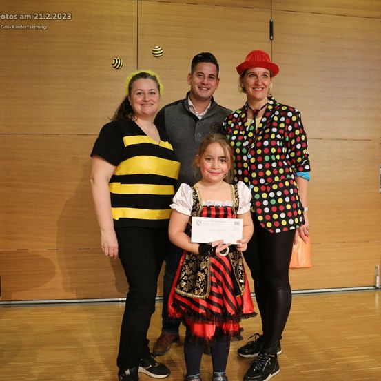 Eine vierköpfige Familie, darunter ein Kind mit einem Zertifikat, steht in einem Raum. Die Frau links trägt ein Bienenkostüm und der Mann in der Mitte einen grauen Pullover. Die Frau rechts trägt ein Punktehemd. Das Datum ist der 21. Februar 2023.