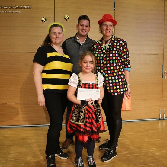 Vier Personen stehen zusammen auf einem Holzboden. Das Mädchen in der Mitte hält ein Zertifikat. Die anderen tragen Kostüme. Hinter ihnen ist eine Holzwand. Im oberen linken Eck steht das Datum 21.2.2023.