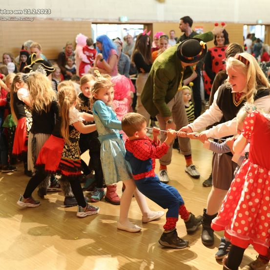 Eine Gruppe von Kindern in Kostümen tanzt in einer Halle, einige als Superhelden verkleidet, ein Kind im Spiderman-Kostüm, mit Erwachsenen um sie herum.