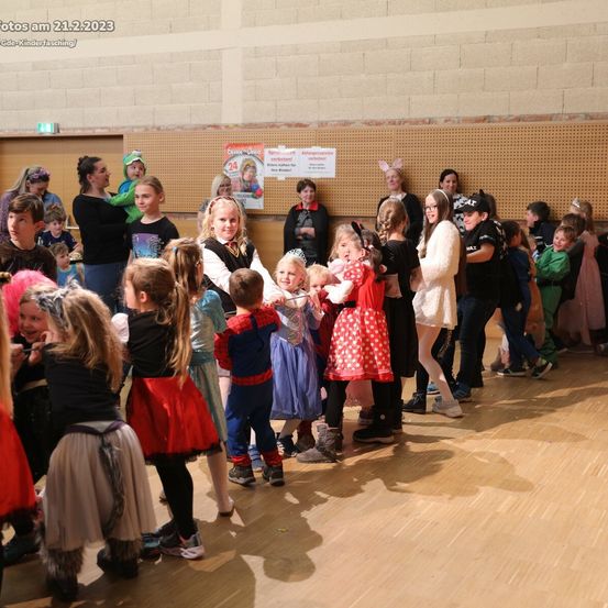 Eine Gruppe von Kindern in verschiedenen Kostümen, einschließlich Superhelden, sind in einem Raum versammelt. Einige Erwachsene stehen hinter ihnen, und ein Poster ist an der Wand. Der Raum hat Holzböden und eine weiße Decke. Das Datum 21.2.2023 ist oben links sichtbar.