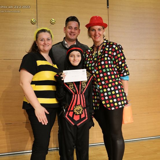 Vier Menschen posieren für ein Foto in einem Raum. Eine Frau trägt ein Bienenkostüm. Ein Mädchen in einem Kostüm hält ein Zertifikat. Die andere Frau trägt ein Punktekleid. Der Mann trägt einen grauen Pullover.