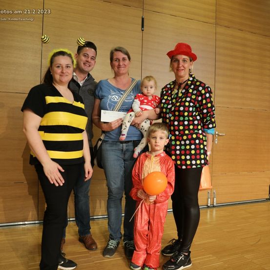 Fünf Personen, alle in Kostümen gekleidet, lächeln und posieren für ein Foto. Einer hält ein Baby, ein anderer ein Zertifikat und ein Kind hält einen Luftballon.