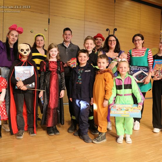 Eine Gruppe von Erwachsenen und Kindern in Kostümen posiert für ein Foto. Die Kinder halten verschiedene Spielzeuge. Die Erwachsenen lächeln und tragen Hüte und Brillen. Ein Banner mit dem Datum '21.2.2023' ist sichtbar.