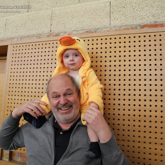 Ein Mann trägt ein Kind auf den Schultern, das in einem gelben Entenkostüm gekleidet ist. Sie lächeln und scheinen für ein Foto zu posieren. Das Bild wurde am 21.2.2023 aufgenommen.