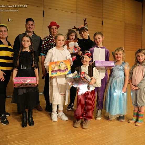 Eine Gruppe von Kindern und Erwachsenen in verschiedenen Kostümen steht zusammen in einem Raum. Einige halten Boxen und Spielzeug. Das Bild ist vom 21.2.2023.