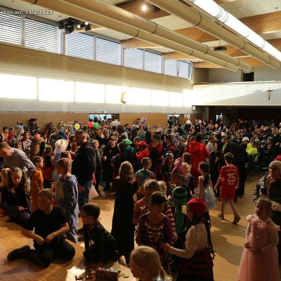 Eine Menschenmenge, hauptsächlich Kinder, ist in einem Gebäude versammelt, wahrscheinlich für eine Halloween-Party. Einige tragen Kostüme, andere halten Luftballons. Der Raum hat Holzböden und -wände mit Deckenleuchten. Es gibt auch Stühle und Tische in dem Bereich.