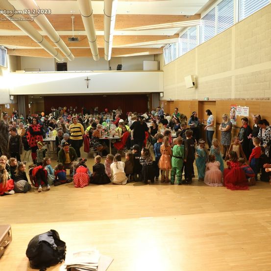 Eine Gruppe von Kindern in Kostümen und Erwachsenen versammelt sich in einem Saal mit Tischen, sitzend und stehend, einige mit Luftballons. Ein Kreuz ist an der Wand sichtbar.