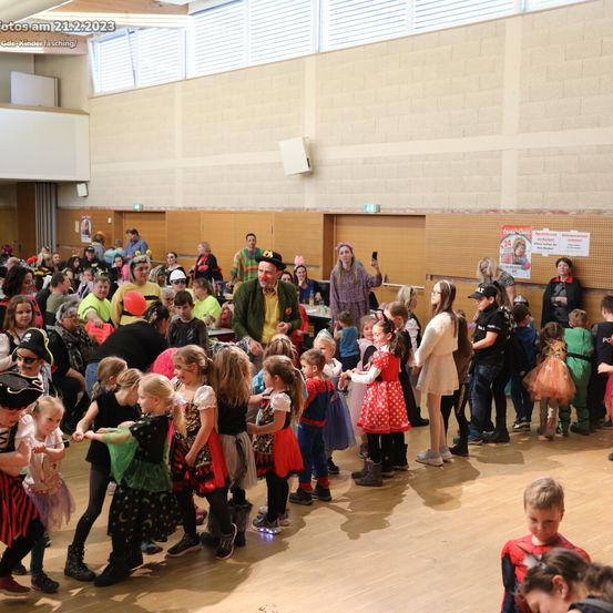 Eine große Gruppe von Kindern und Erwachsenen in verschiedenen Kostümen versammelt sich in einem Saal, wahrscheinlich für eine Halloween-Veranstaltung. Einige stehen in einer Schlange, während andere interagieren. Eine Person in der Mitte hält ein Handy. Der Raum hat Holzböden und weiße Wände mit gerahmten Bildern.
