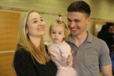 Eine lächelnde Familie mit einem kleinen Mädchen in einem rosa Kleid und einer rosa Schleife, das in einem Raum mit einer Holzwand und einer Person im Hintergrund steht.