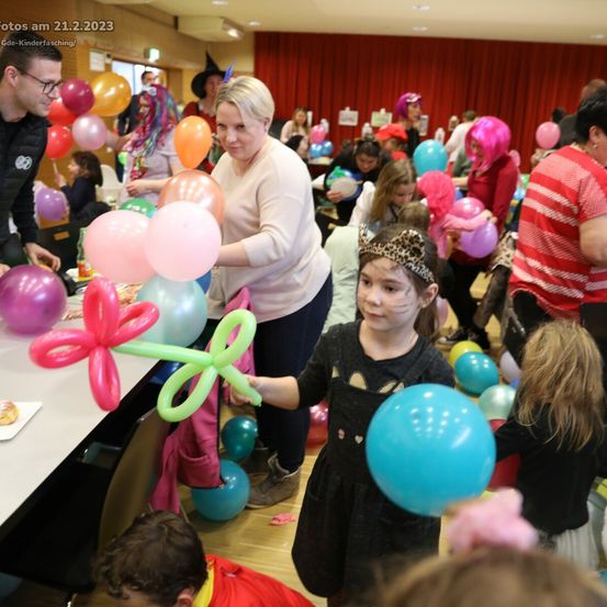 Eine farbenfrohe Veranstaltung mit Erwachsenen und Kindern. Ein Mädchen, das eine Ballonblume hält, steht im Vordergrund. Es gibt viele Luftballons in der Luft.