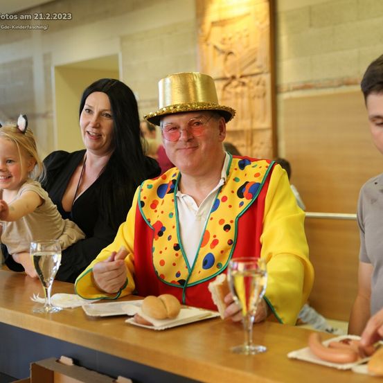 Eine Familie mit einem Kind in einem Clownkostüm sitzt an einem Tisch mit Essen und Getränken.