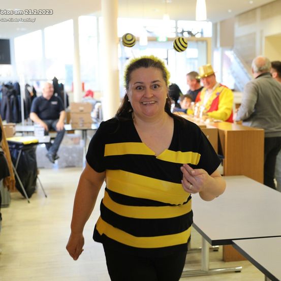 Eine Frau in einem Bienenkostüm geht durch einen Raum mit anderen Menschen und Tischen. Ein Datumswasserzeichen ist oben links.