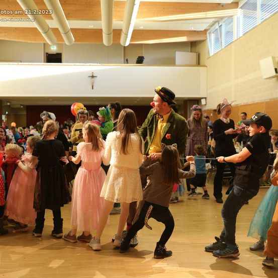 Eine Gruppe von Kindern in Kostümen tanzt mit einem Clown und einem Künstler in einem großen Saal, mit Erwachsenen und weiteren Kindern im Hintergrund.