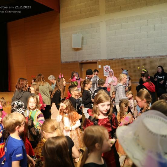 Eine große Gruppe von Kindern in verschiedenen Kostümen versammelt sich in einer Halle. Viele tragen Masken, Hüte und halten Requisiten. Im Hintergrund befinden sich eine Bühne und Wände mit einem Schild.