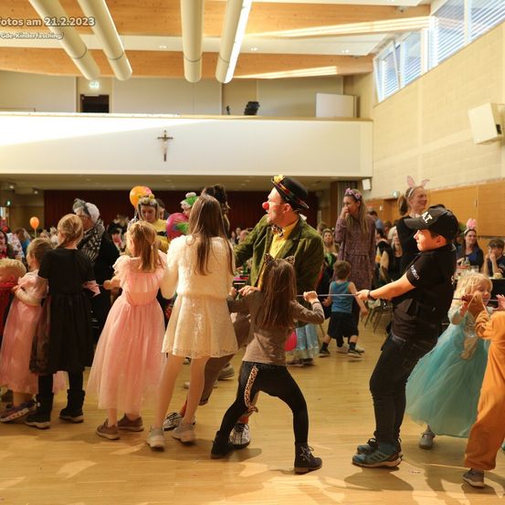 Eine große Gruppe von Kindern und Erwachsenen in Kostümen tanzt am 21.2.2023 in einem Saal. Einige Kinder tragen Hasenohren, andere Clowns-Make-up. Ein Kind hält einen Stock.