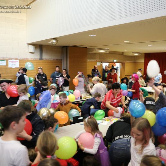 Eine große Gruppe von Menschen, darunter Kinder und Erwachsene, versammelt sich in einem Raum. Sie halten Luftballons und scheinen ein Ereignis zu feiern. Tische mit Essen und anderen Gegenständen sind aufgebaut. Das Datum 21.2.2023 ist in der oberen linken Ecke sichtbar.