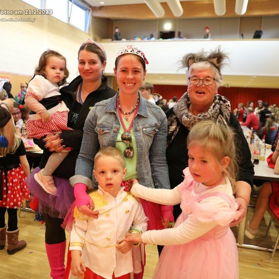 Eine vierköpfige Familie posiert für ein Foto bei einer festlichen Veranstaltung am 21. Februar 2023, mit Kindern in Kostümen und lächelnden Erwachsenen, umgeben von anderen Gästen und einem dekorierten Raum.
