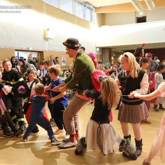 Eine Gruppe von Kindern in Kostümen und ein Mann in einem Hut spielen auf einer Tanzfläche. Das Datum des Fotos ist 21.2.2023.