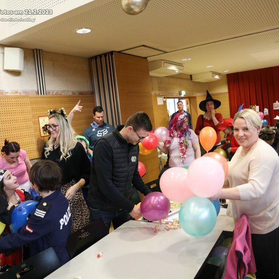 Eine Gruppe von Menschen in einem Raum, einige in Kostümen gekleidet, versammeln sich um einen Tisch voller Luftballons. Eine Frau auf der rechten Seite hält einen Luftballon. Das Bild ist vom 21.2.2023.