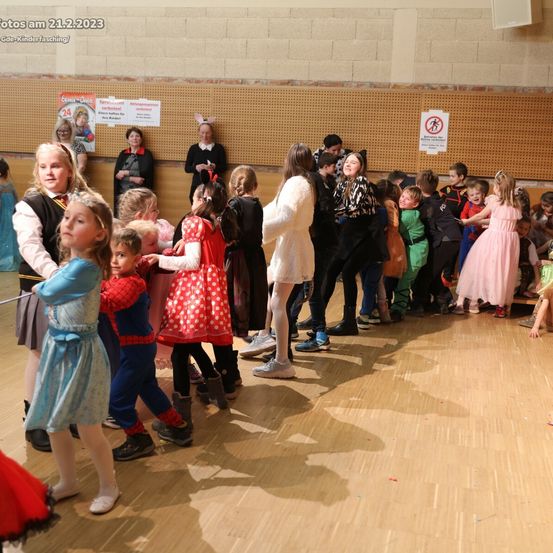 Kinder in Kostümen versammeln sich in einem Saal, einige halten Händchen und spielen. Ein Schild an der Wand zeigt 'Kein Gehen'. Das Datum 21.2.2023 ist oben links sichtbar.